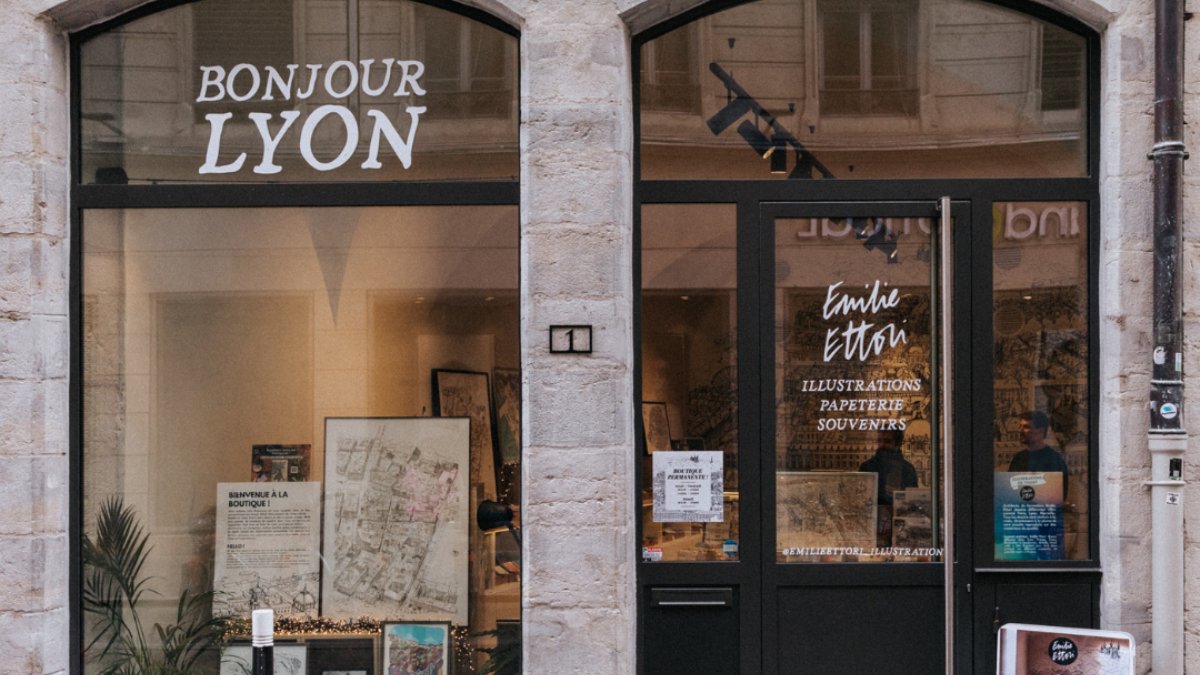La vitrine de la boutique Émilie Ettori Illustration, avec l’enseigne « Bonjour Lyon » et des affiches exposées derrière la porte vitrée, à Lyon La vitrine de la boutique Émilie Ettori Illustration, avec l’enseigne « Bonjour Lyon » et des affiches exposées derrière la porte vitrée, à Lyon