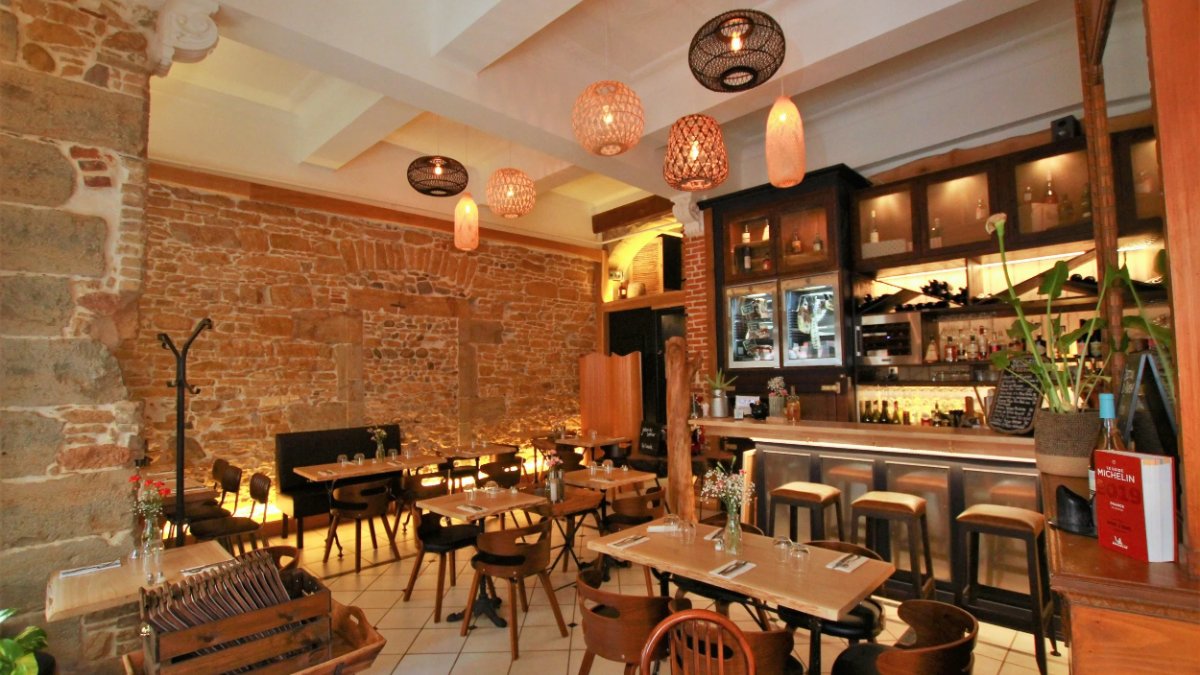 La salle de L’Arbre Sec à Lyon, murs de pierre, tables en bois et comptoir de bar éclairé en fond La salle de L’Arbre Sec à Lyon, murs de pierre, tables en bois et comptoir de bar éclairé en fond