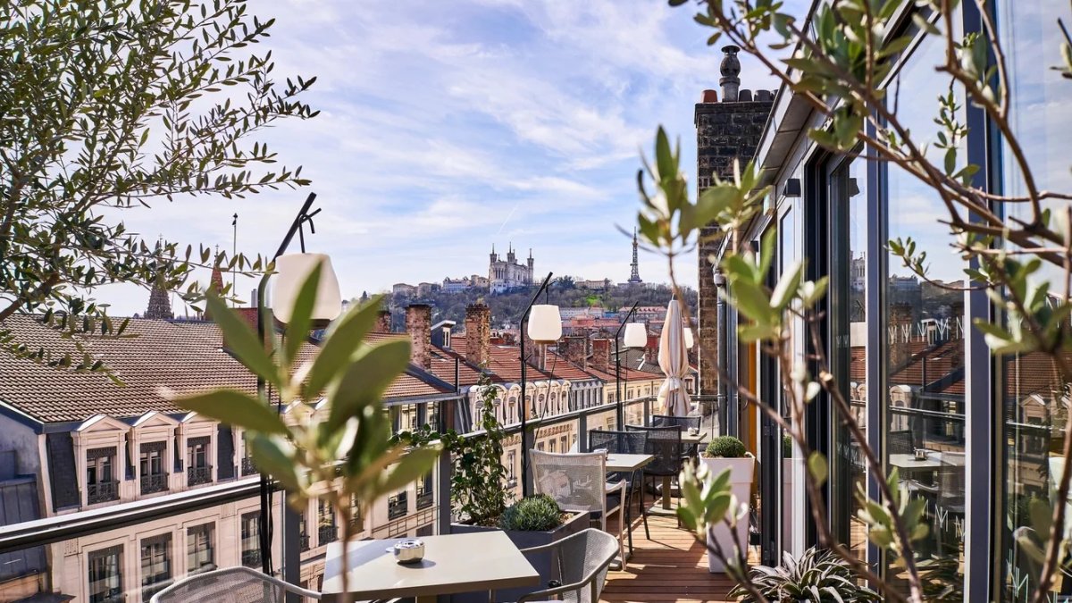 Une terrasse sur les toits de Lyon avec tables, plantes et vue sur les toits et la basilique de Fourvière, au Rooftop de la Maison Nô