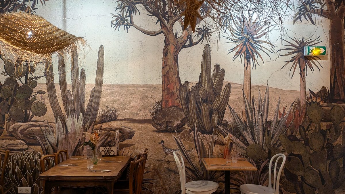 La salle de Maison Courgette à Lyon, avec ses tables en bois, ses chaises bistrot et sa grande fresque murale de cactus et de plantes du désert La salle de Maison Courgette à Lyon, avec ses tables en bois, ses chaises bistrot et sa grande fresque murale de cactus et de plantes du désert