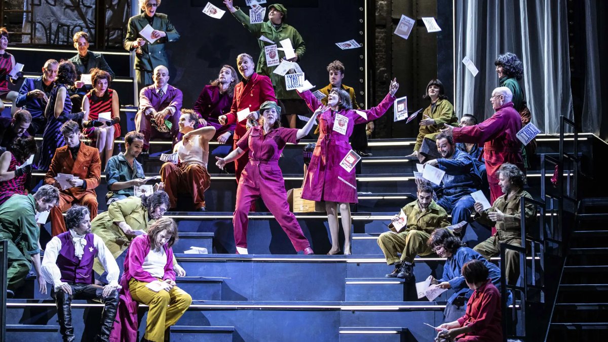 Des chanteurs en costumes colorés assis sur des gradins sur la scène de l’Opéra national de Lyon, pendant que des feuilles de papier volent dans l’air