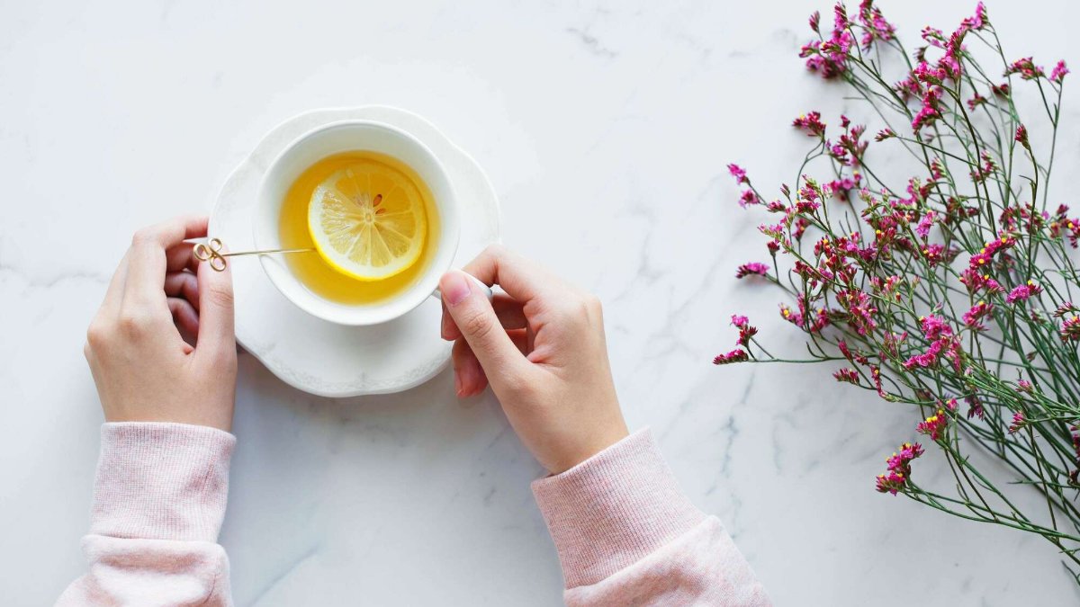 Deux mains tenant une tasse de tisane au citron, à côté d’un bouquet de petites fleurs roses, sur une table en marbre blanc Deux mains tenant une tasse de tisane au citron, à côté d’un bouquet de petites fleurs roses, sur une table en marbre blanc