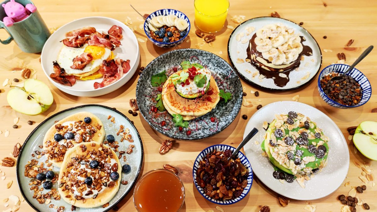 Une table en bois garnie de pancakes aux myrtilles, à la banane et au chocolat, entourés de pommes et de petits bols chez Yummy Pancakes à Lyon